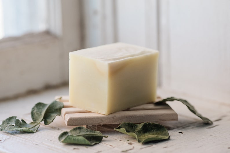 A block of cream-colored solid dish soap lays on a slatted wooden tray. Dried makrut lime leaves are scattered on top and around the soap. Soft morning light from the adjacent window illuminates the soap from the left side.
