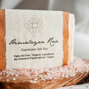 Op de afbeelding: Een witte reep handgemaakte Himalayazoutzeep van Himalayan Rove met een bruin papieren etiket. Het etiket heeft een decoratief bloemmotief en de tekst "Himalayan Rove Handmade Salt Bar Palm-Oil Free Organic Ingredients No Chemical Preservatives Vegan". De zeepreep ligt op een houten oppervlak met roze Himalaya-zoutkristallen.