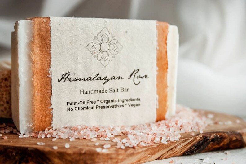 Op de afbeelding: Een witte reep handgemaakte Himalayazoutzeep van Himalayan Rove met een bruin papieren etiket. Het etiket heeft een decoratief bloemmotief en de tekst "Himalayan Rove Handmade Salt Bar Palm-Oil Free Organic Ingredients No Chemical Preservatives Vegan". De zeepreep ligt op een houten oppervlak met roze Himalaya-zoutkristallen.
