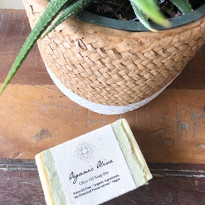 A cream-colored soap bar wrapped in sage and off-white flower-seeded paper sits next to a succulent plant in a woven pot. Both are on a wooden table. The soaps label reads, "Organic Olive Soap Bar," along with a brief description.