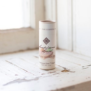 May include: A white cardboard tube with a brown label. The label has the text "Sandalwood Spice Organic Deodorant" and "Aluminum Free * Vegan" with a net weight of 2 oz. The label also features a floral design and a picture of a sandalwood stick.