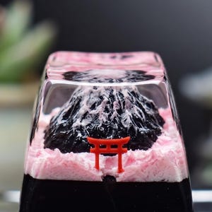 May include: A clear resin keycap with a miniature black mountain and white snowy peak, set against a pink and black base. A small red torii gate is at the mountain's base, creating a Japanese theme.