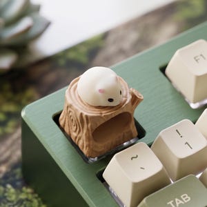 Puede incluir: Una tecla personalizada con una figura de ratón blanco sobre un pequeño tocón de árbol. La tecla está diseñada para un teclado mecánico, con una base verde y teclas blanquecinas visibles en el fondo.