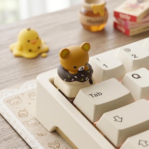 May include: A cream-coloured keyboard with white keycaps. A brown bear figurine sits on a keycap, atop a chocolate donut with sprinkles. A yellow chick figurine is also visible. A small jar of honey and a stack of books are in the background.