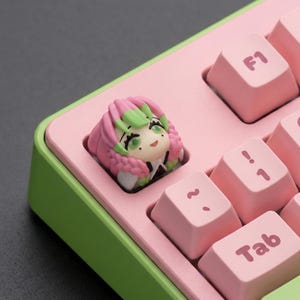 May include: A custom keyboard keycap featuring a character with pink and green hair, set on a pink keyboard with green trim. The keycaps are pink with white lettering. The keycap is a unique accessory for mechanical keyboards.