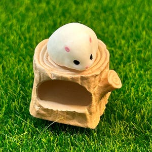 May include: A small white hamster figurine with pink ears and black eyes is sitting on a brown wooden log with a hollowed-out space. The log is on a green artificial grass surface.