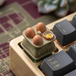 May include: A novelty keyboard keycap shaped like a miniature egg box, with four brown eggs, one cracked to show a yellow yolk. The keycap is olive green and says "BAD EGG". The keyboard has black keys and a brass frame.