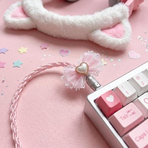 May include: A braided light pink USB cable featuring a decorative light pink lace bow with a central pearl-like heart charm, connected to a mechanical keyboard with pink and white keycaps, including one keycap with a white heart icon.