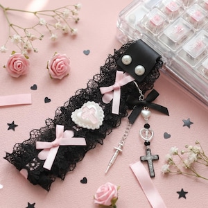 Peut inclure: Un tour de cou en dentelle noire orné de deux nœuds en ruban rose pâle, d'un cœur blanc avec une croix rose, et d'une chaîne argentée avec un pendentif croix noire, un petit cœur et une breloque seringue, fixé par deux perles blanches.