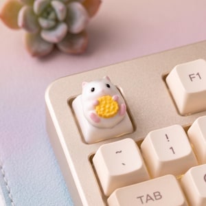 May include: A white and pink hamster-shaped keycap holding a yellow cookie, installed on a beige keyboard. The keycap replaces a standard key. The keyboard has white keys with black lettering.
