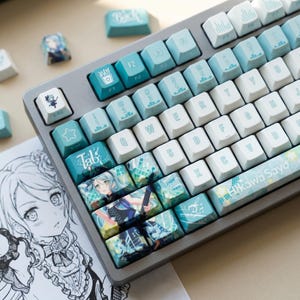 Può includere: Una tastiera meccanica grigia con tasti personalizzati nei toni del blu e del bianco. Alcuni tasti presentano illustrazioni in stile anime. La tastiera è su una superficie bianca con un disegno di un personaggio anime.