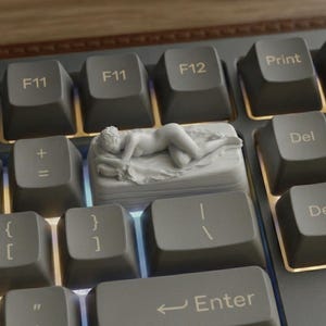 Puede incluir: Una tecla personalizada para teclado mecánico con una escultura blanca de una figura reclinada sobre una cama. La tecla está colocada entre las teclas F11, F12 e Impr Pant. Las teclas del teclado son grises.