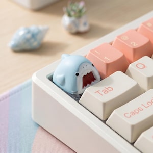 May include: A light blue shark-shaped artisan keycap with a wide-open mouth and red interior, positioned on a white mechanical keyboard next to cream-colored keys with pink lettering, including the Tab and Caps Lock keys.