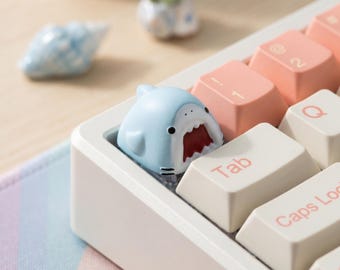 May include: A light blue shark-shaped artisan keycap with a wide-open mouth and red interior, positioned on a white mechanical keyboard next to cream-colored keys with pink lettering, including the Tab and Caps Lock keys.