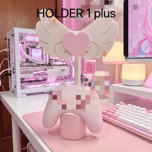 Peut inclure: Un support de manette de jeu rose avec un cœur central entouré de deux ailes blanches. Le support possède une base cylindrique et un montant vertical. Le texte "HOLDER 1 plus" est imprimé en noir en haut de l'image.