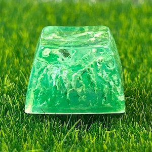 May include: A green and white resin keycap with a swirling, marbled pattern. The keycap is sitting on a bed of green grass.