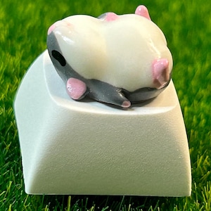 May include: A small, white and gray hamster figurine with pink ears and nose, lying on its back on a white keycap.