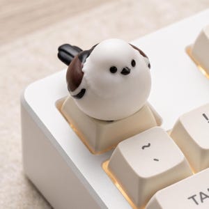 Puede incluir: Una tecla con forma de pájaro blanco y marrón se asienta sobre una tecla de teclado mecánico de color crema. La figura del pájaro tiene una cabeza blanca redonda, ojos negros, un pequeño pico negro y detalles de alas en marrón y negro.