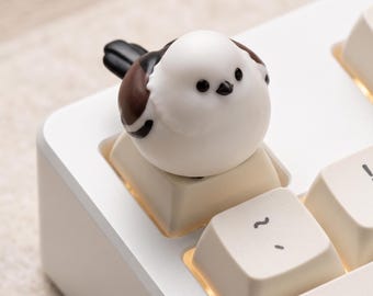 May include: A white and brown bird-shaped keycap sits atop a cream-colored mechanical keyboard key. The bird figure has a round white head, black eyes, a small black beak, and brown and black wing details, replacing a standard keyboard key.