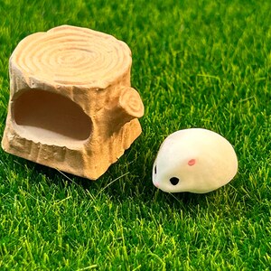 May include: A small white hamster figurine with pink ears and black eyes, sitting on a green artificial grass surface next to a brown wooden log with a hollowed-out space.