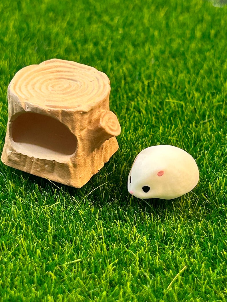 May include: A small white hamster figurine with pink ears and black eyes, sitting on a green artificial grass surface next to a brown wooden log with a hollowed-out space.