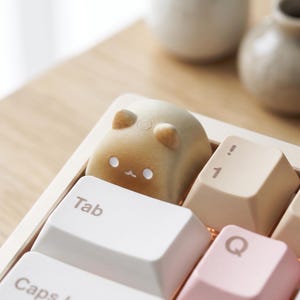 May include: A custom mechanical keyboard keycap shaped like a small, tan, rounded creature with two ears, two white circular eyes, and a tiny mouth. The keycap is positioned on a keyboard next to a white Tab key and a beige key with the number 1.