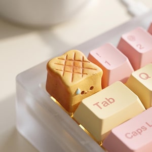 Peut inclure: Une touche de clavier artisanale en forme de tranche de pain grillé avec un motif quadrillé sur le dessus et une petite larme bleue sur le côté. La touche est installée sur un clavier mécanique à côté de touches jaune clair et roses.