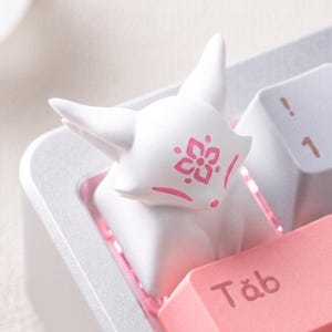 May include: A white fox-shaped keycap with pink floral details is placed on a keyboard. The keycap features a pink flower design on its forehead and pink markings. The keyboard keys are white and pink.