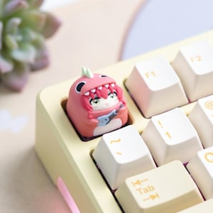 May include: A custom keyboard keycap featuring a pink dinosaur design with a cartoon character holding a guitar. The keycap is set on a light yellow keyboard with white keys. The keycap is a unique accessory for mechanical keyboards.
