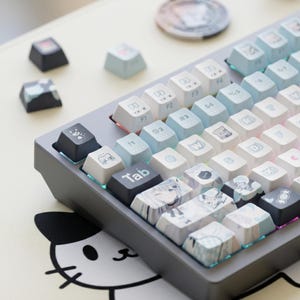 Può includere: Una tastiera meccanica grigia con tasti personalizzati. I tasti sono un mix di bianco, azzurro e nero, con varie icone e illustrazioni in stile anime. Alcuni tasti sono sparsi su un tappetino da scrivania a tema gatto.