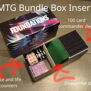 Può includere: Un inserto per scatola MTG Bundle con il testo "MTG Bundle Box Insert" e "FOUNDATIONS". La scatola contiene un mazzo comandante da 100 carte, dadi, contatori vita e bustine per carte draft.