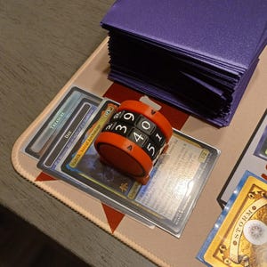 May include: An orange and black dice counter sits atop a stack of trading cards. The counter has numbers 0-9 and is used to track life points in a card game. A stack of purple card sleeves is in the background.