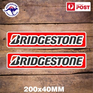Puede incluir: Dos pegatinas rojas y blancas con la palabra "BRIDGESTONE" en letras negras. Las pegatinas miden 200 mm x 40 mm.