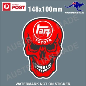 Puede incluir: Pegatina de calavera roja con un logotipo de Toyota blanco dentro de un círculo. La calavera tiene ojos negros y una sonrisa blanca. La pegatina mide 148 mm x 100 mm.