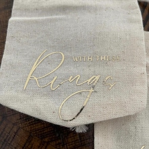 May include: A white fabric ring bearer bag with gold lettering that says "With these rings".