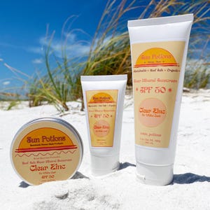 May include: Three bottles of Sun Potions Clear Zinc SPF 50 sunscreen. The bottles are white with orange and yellow labels. The labels say "Sun Potions", "Clear Zinc", "SPF 50", and "Reef Safe Mineral Sunscreen". The bottles are on a white sandy beach.