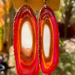 May include: A pair of statement earrings with a red, orange, and white agate design. The earrings are oval shaped and have a gold-colored metal backing.