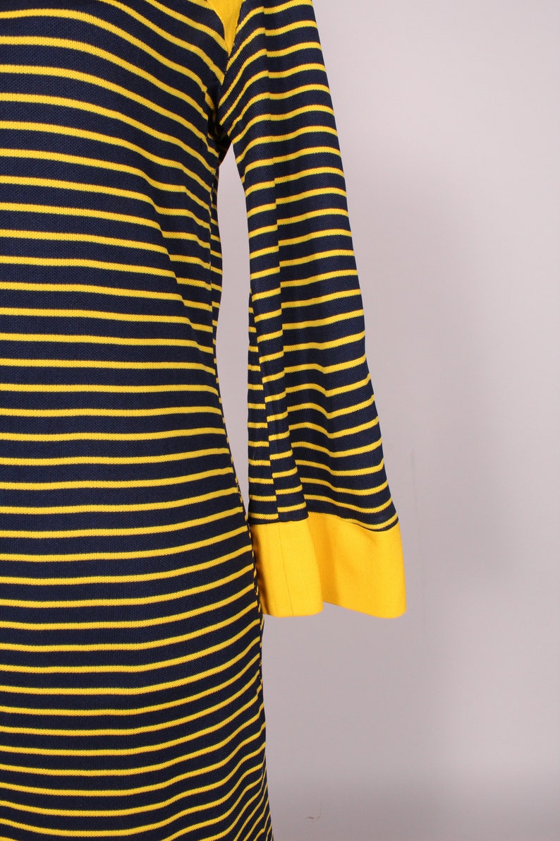 Puede incluir: Un vestido de manga larga con rayas amarillas y azul marino. El vestido tiene una manga acampanada y un pu&ntilde;o amarillo.