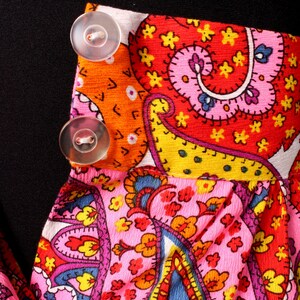 May include: A pink and orange paisley patterned fabric with two white buttons. The fabric has a red, yellow, and blue paisley design.