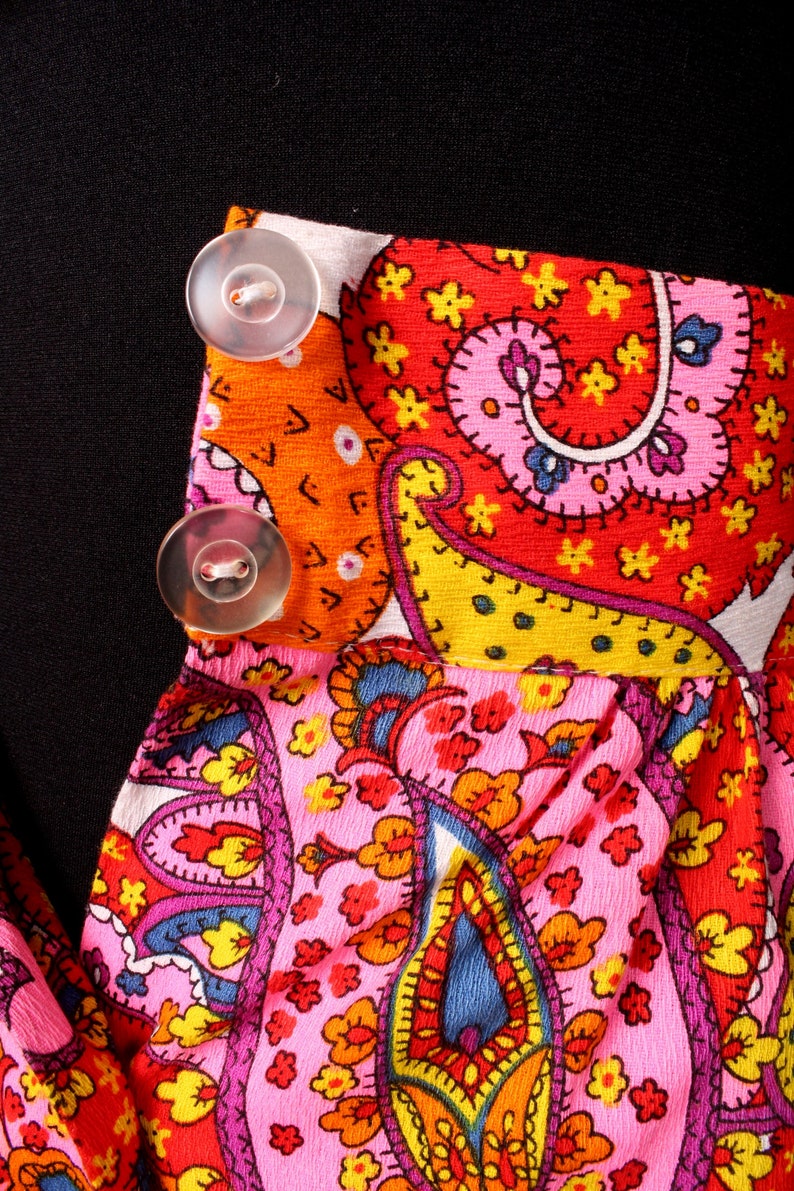 May include: A pink and orange paisley patterned fabric with two white buttons. The fabric has a red, yellow, and blue paisley design.