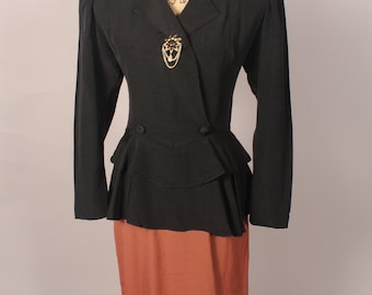 May include: A black dress with a peplum waist and a long sleeve jacket. The jacket has a gold brooch and a double breasted closure. The dress is a two-piece set.