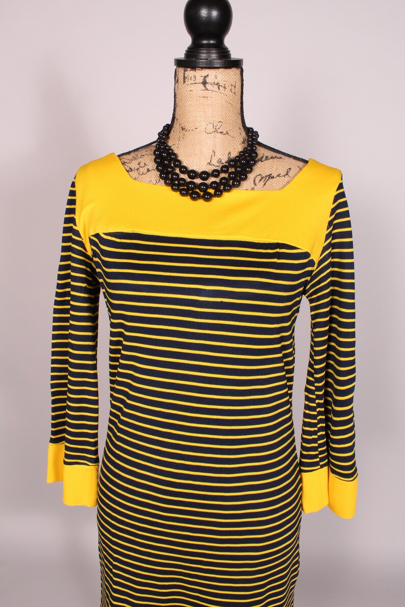 Puede incluir: Un vestido de rayas amarillas y negras con un escote amarillo y mangas largas. El vestido tiene un collar de cuentas negras.