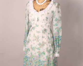 Puede incluir: Un vestido floral blanco vintage con mangas largas, cuello con volantes y dobladillo de encaje. El vestido presenta un delicado estampado de flores azules, rosas y verdes. Un collar de cuentas blancas se exhibe en un maniquí.