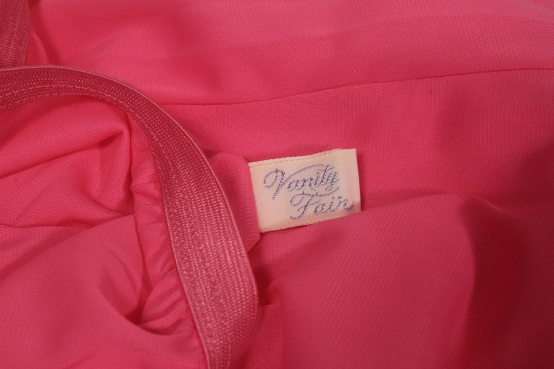 May include: A close-up of a pink fabric garment with a white label that says "Vanity Fair" in blue lettering.