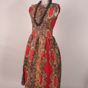 以下が含まれることがあります： 赤、茶色、金色の幾何学模様の袖なしドレス。ドレスは体にフィットしたボディとふっくらとしたスカート。ドレスはマネキンに着せられており、黒いビーズのネックレスをしています。