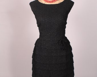 May include: A black dress with a ruffled skirt and a small flower detail at the waist. The dress is on a mannequin and has a coral necklace.