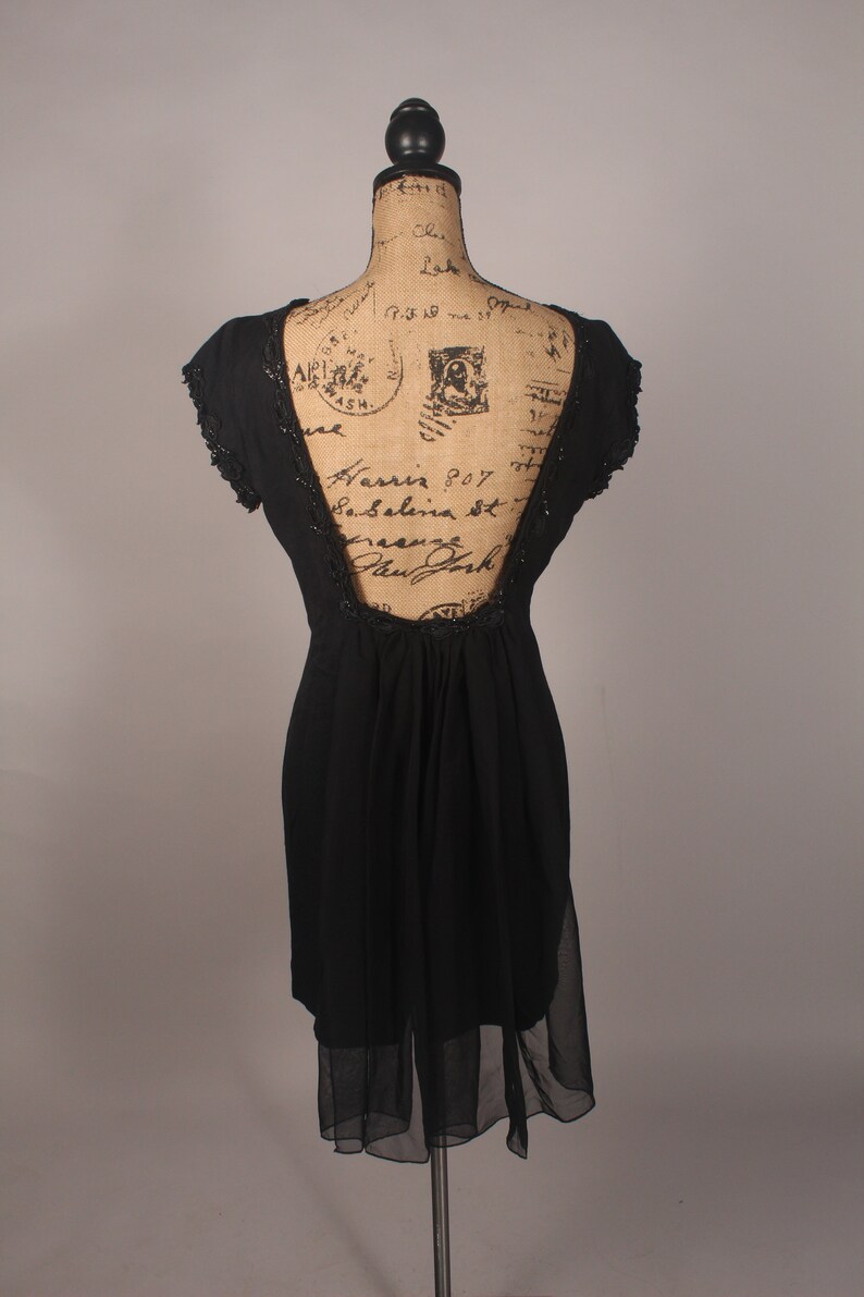 May include: A black sleeveless dress with a sheer overlay and a low back. The dress is on a dress form with a vintage-style pattern.