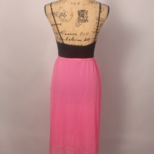 May include: A pink maxi slip skirt with a lace trim. The skirt is on a mannequin and is made of a silky material.