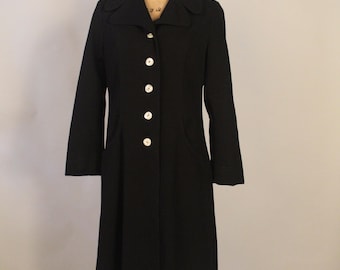 May include: A black coat with a classic design, featuring a collar, long sleeves, and a row of white buttons down the front. The coat has a tailored fit and appears to be made of a smooth fabric. It is displayed on a mannequin.