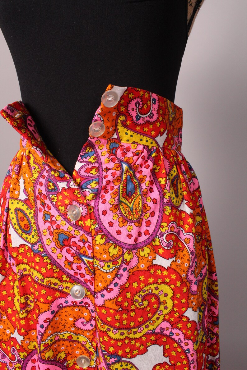 May include: A white skirt with a colorful paisley pattern in shades of pink, orange, yellow, and blue. The skirt has a button-up front closure.